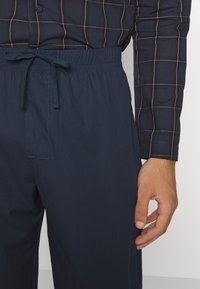Pantalones azul marino con cintura elástica y cordón, con una textura suave y un bolsillo lateral, combinados con una camisa a cuadros.