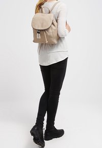 Femme portant un sac à dos beige, un haut blanc à manches longues superposées, un pantalon skinny noir et des bottes noires, se tenant de dos.
