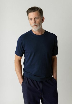 Uomo con capelli grigi e barba, in piedi con le mani nelle tasche, che indossa una camicia blu navy a maniche corte e pantaloni scuri davanti a uno sfondo semplice.