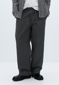 Grå, lette bomuldsbukser med elastisk talje og snøre. Fladt forstykke, lige ben og manchetter, der sidder over sorte støvler.