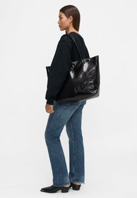 Borsa tote in pelle nera con una finitura lucida, dotata di due manici e un design spazioso, accompagnata da un modello in abbigliamento casual.