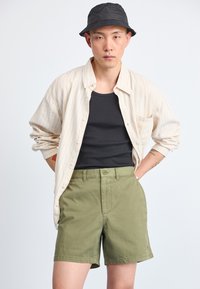 Ανοιχτόχρωμο πουκάμισο με ρίγες, φορεμένο πάνω από μαύρο τοπ, συνδυασμένο με ελιόχρωμες βερμούδες και μαύρο καπέλο μπέιζμπολ. Ομαλές υφές υφασμάτων ορατές.