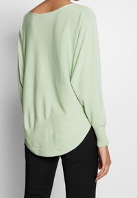 Suéter suelto de color verde claro, con un escote amplio y mangas largas. La tela parece suave, con una textura acanalada y dobladillo redondeado.