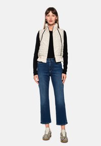 Gesteppte beige Weste mit hohem Kragen, schwarzes Langarmshirt und blaue, abgeschnittene Jeans. Die Schuhe haben ein Muster in neutralen Farbtönen.