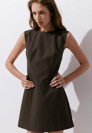 Mujer con cabello ondulado que lleva un vestido sin mangas a cuadros de color marrón oscuro, de pie con las manos en las caderas contra un fondo liso y claro.