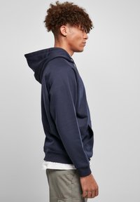 Marineblå hettegenser laget av myk stoff, med en frontlomme, ribbestrikkede mansjetter og en justerbar snorhette. Glatt tekstur.