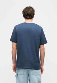 Άνδρας με κοντά καστανά μαλλιά που φοράει απλή μπλε μπλούζα και ανοιχτόχρωμα τζιν σορτς τύπου cargo, φαίνεται από πίσω.