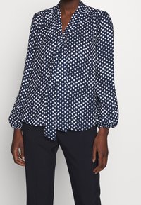 Blusa blu navy con stampa di cuori bianchi; presenta un collo annodato e maniche a palloncino, realizzata in tessuto leggero, abbinata a pantaloni neri.