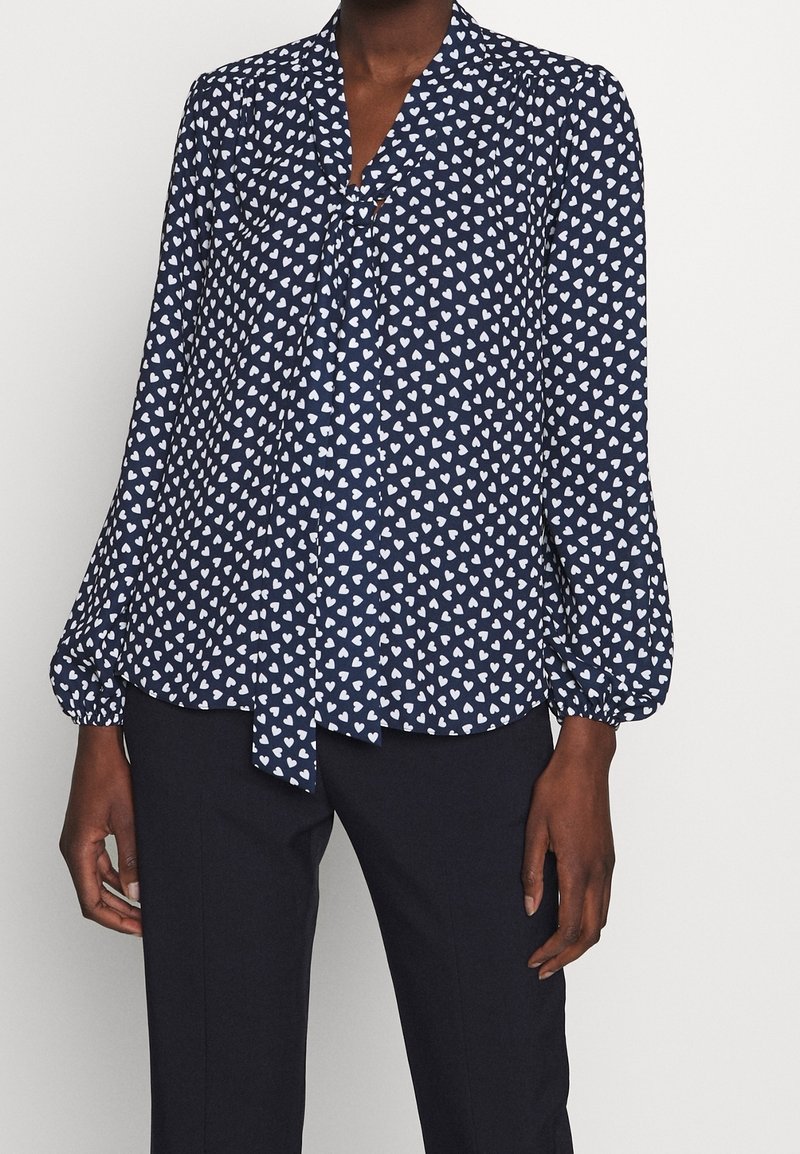 Blusa blu navy con stampa di cuori bianchi; presenta un collo annodato e maniche a palloncino, realizzata in tessuto leggero, abbinata a pantaloni neri.