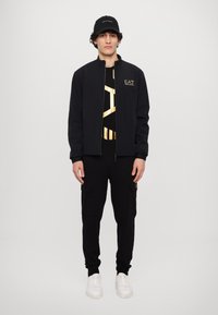 Svart zip-up jacka, svart t-shirt med guldmönster, svarta cargo-byxor och vita sneakers. En hatt med logotyp fulländar outfiten.