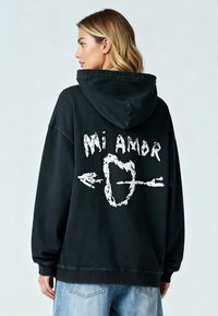 Haut à capuche surdimensionné noir avec une impression graphique blanche au dos. Comprend une capuche avec cordon de serrage et des poignets côtelés.