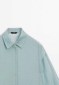 Chemise bleu clair à manches longues avec col pointu, tissu texturé et boutons visibles sur le devant, posée à plat sur un fond blanc.