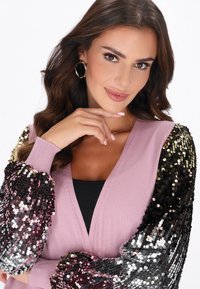 Różowy kardigan z czarnymi i iryzującymi rękawami ozdobionymi cekinami. Posiada dekolt w serek, ściągacze przy rękawach i dopasowany fason.