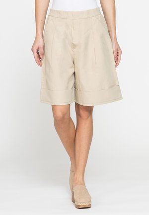 Donna che indossa pantaloncini beige larghi fino al ginocchio, top bianco e scarpe beige con tacco slip-on su sfondo uniforme.