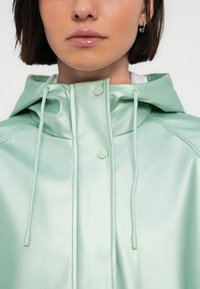 Ανοιχτό πράσινο αδιάβροχο μπουφάν με λεία, γυαλιστερή υφή, ρυθμιζόμενα κορδόνια και κουμπώματα με κουμπιά στο λαιμό.
