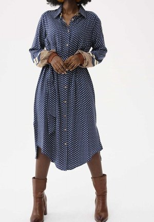 Frau in einem blau gemusterten Midikleid mit Knopfleiste und gemusterten Bündchen, stehend in braunen Stiefeletten auf weißem Hintergrund.