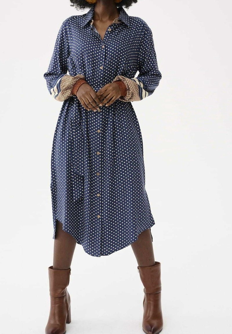Femme en robe midi bleue à motifs avec boutons et poignets à motifs mélangés, debout avec des bottines marron à talons sur fond blanc.