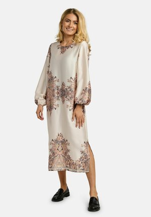 Kobieta o blond włosach, ubrana w długą beżową sukienkę w różowe i czarne kwiatowe wzory oraz czarne mokasyny, uśmiechająca się i stojąca.