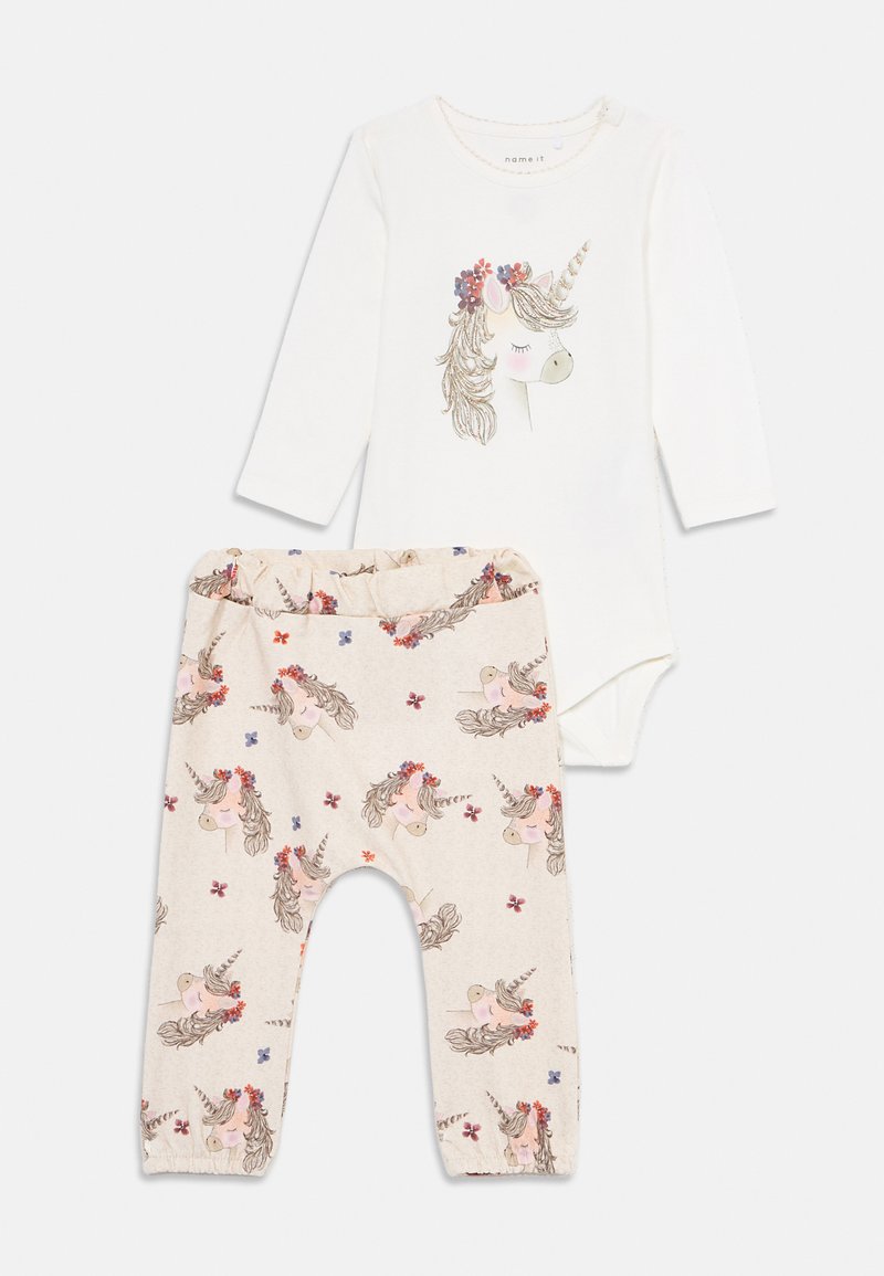 Weißer Babybody mit langen Ärmeln, der ein Einhorn-Gesicht und einen Blumenkronen-Aufdruck zeigt, kombiniert mit beigen Hosen, die ein passendes Einhorn- und Blumenmuster aufweisen.