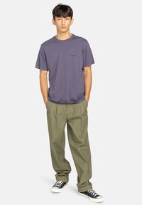 T-shirt di cotone viola con scollatura rotonda e logo discreto, abbinata a pantaloni chinos larghi color verde oliva e sneakers nere. Design semplice e casual.