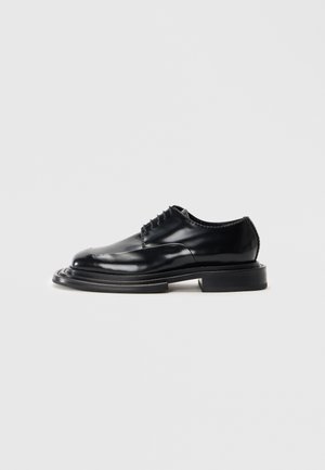 Chaussure en cuir noir avec une finition brillante, bout rond, lacets et une semelle épaisse et plate. Présente un design sculpté en couches.