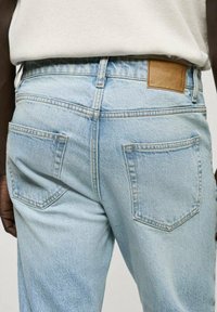 Jeans din denim de un albastru deschis, cu o croială dreaptă, având două buzunare pe spate și o etichetă din piele maro pe talie. Textură moale, design casual.