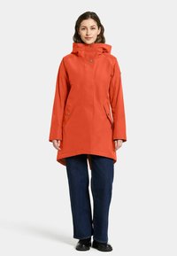 Vandtæt orange parka med hætte, lange ærmer, sidelommer og en let længere ryg. Sammen med mørkeblå vide jeans.