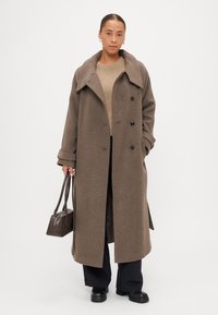 Brūns vilnas mētelis ar plašu apkakli, dubultā pogu aizdare un pārāk liels piegriezums. Stilizēts ar beigu džemperi un melniem biksēm.