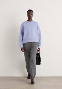 Lyselilla oversized genser, grå joggebukser og svart håndveske. Modellen har på seg mørke sko; den enkle bakgrunnen fremhever klærne.