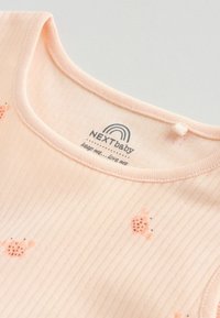 Gros plan sur un body bébé rose pâle côtelé avec de petits imprimés de coccinelles orange et une étiquette NEXTbaby à l'intérieur du col.