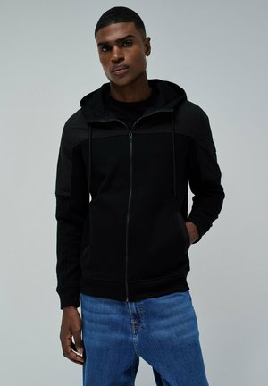 Sudadera negra con cremallera en la parte delantera, cordones y dos bolsillos laterales. Presenta una textura de tela suave con un diseño ajustado. Se lleva con jeans azules.