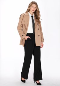 Bézs ballonkabát dupla soros gombolással, nagy gombokkal és vállpántokkal, fekete bő szárú nadrággal és fehér inggel párosítva.