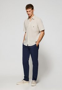 Lichtbeige korte mouw shirt met een borstzak en een klein logo, gecombineerd met marineblauwe broeken en lichtbeige casual sneakers.