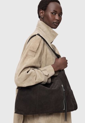 Isik, kes kannab beežist seemisnahast trentšmantlit ja hoiab suurt tumepruuni seemisnahast käekotti, millel on külje peal tõmblukuga tasku.