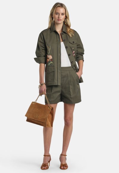 Woman wearing olive green floral-embroidered jacket and matching shorts, white top, brown handbag with gold chain, and brown high-heeled sandals.