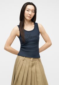 Jeune femme aux longs cheveux foncés portant un haut sans manches bleu marine avec le texte « Guess » et une jupe plissée beige, debout les mains derrière le dos.