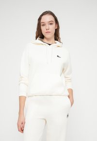 Sweat à capuche couleur crème avec une poche avant et une capuche ajustable à cordon, fabriqué en tissu doux. Dispose d'un petit logo noir sur la poitrine.