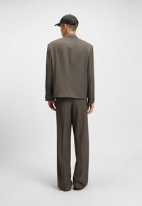 Brown textured suit with relaxed fit, featuring a single back vent. Paired with a black cap. Focus on tailored design and contemporary silhouette.