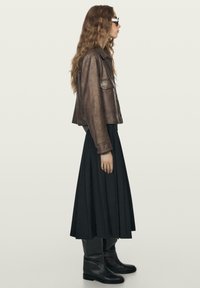 Žena v profilu, oblečená v hnědé kožené bundě, černé skládací midi sukni, černých vysokých botách a černých slunečních brýlích na jednobarevném pozadí.