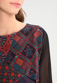 Top multicolore con motivo geometrico in rosso, blu e verde su uno sfondo scuro, con maniche nere trasparenti.
