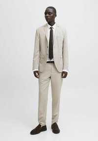Completo beige con giacca su misura e pantaloni a gamba dritta, abbinato a una camicia bianca e una cravatta marrone scuro. Scarpe marrone scuro completano l'outfit.