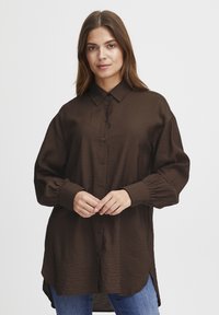 Brown long-sleeve button-up shirt with a relaxed fit, textured fabric, and adjustable cuffs, paired with blue jeans. Side slits at the hem.