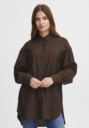 Brown long-sleeve button-up shirt with a relaxed fit, textured fabric, and adjustable cuffs, paired with blue jeans. Side slits at the hem.
