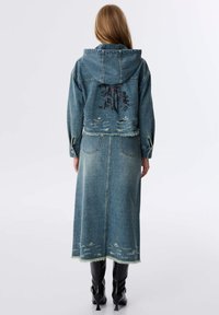 Jeansjacke mit Kapuze, die ein Grafikdesign mit einem schwarzen Wald auf dem Rücken zeigt, mit abgenutzten Details, und einem langen Jeansrock mit franslichem Saum.