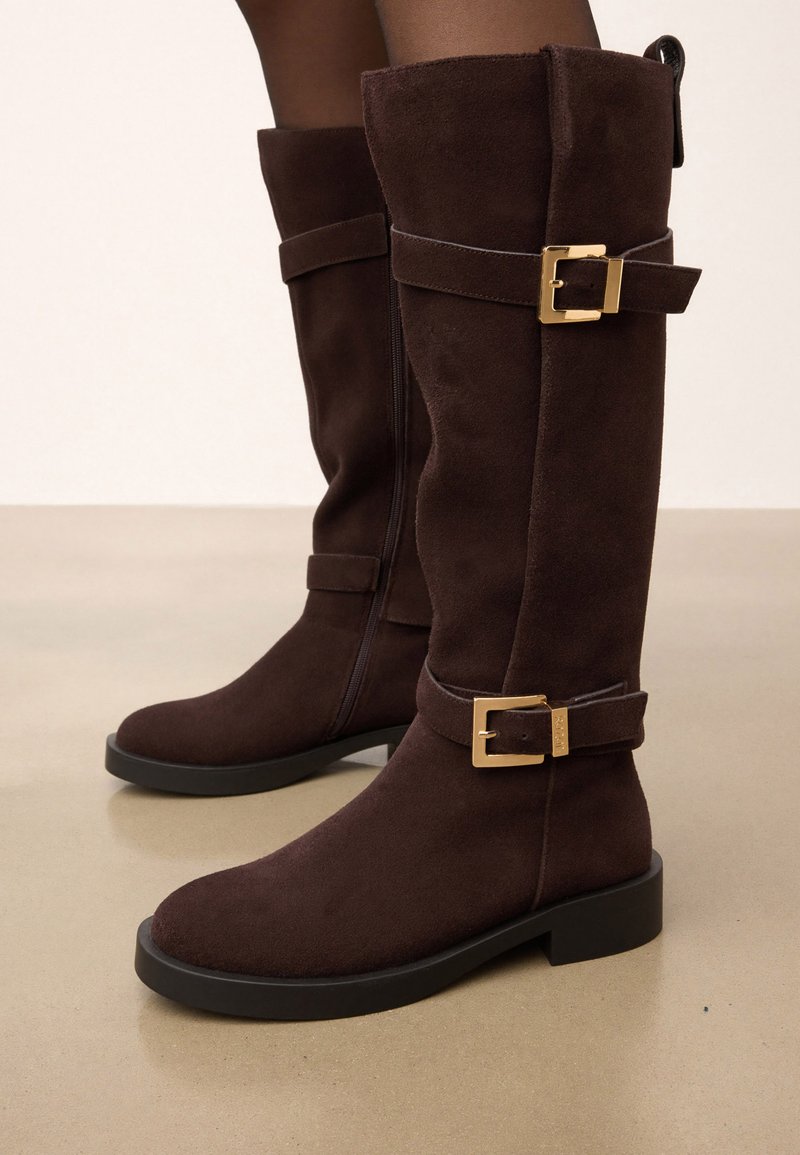 Tumši brūni zamšas ceļgala augstuma zābaki ar melniem zoleņiem un zelta sprādzēm, valkāti uz kājām uz gaiša fona.