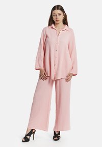Lichtroze, gestructureerde katoenen button-up shirt met lange mouwen en een kraag, gecombineerd met wijde broek in hetzelfde materiaal. Zwarte hoge hakken.