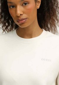 Felpa bianca girocollo in misto cotone, con il logo "GUESS" in rilievo sulla parte sinistra del petto e una finitura liscia e testurizzata.