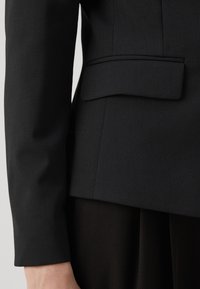 Zwart getailleerd blazer met een gestructureerd ontwerp, met een gladde textuur en een platte zakdetail aan de linkerkant.