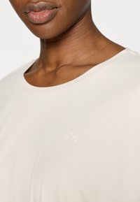 Μπεζ κοντομάνικο μπλουζάκι με λεία υφή και στρογγυλή λαιμόκοψη· διαθέτει μικρό, ανο ιχτόχρωμο λογότυπο στην αριστερή πλευρά κοντά στο στήθος.