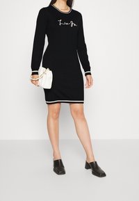 Zwarte gebreide jurk met lange mouwen, witte gestreepte accenten bij de manchetten en zoom, met geborduurd logo op de borst. Model houdt een witte handtas vast.